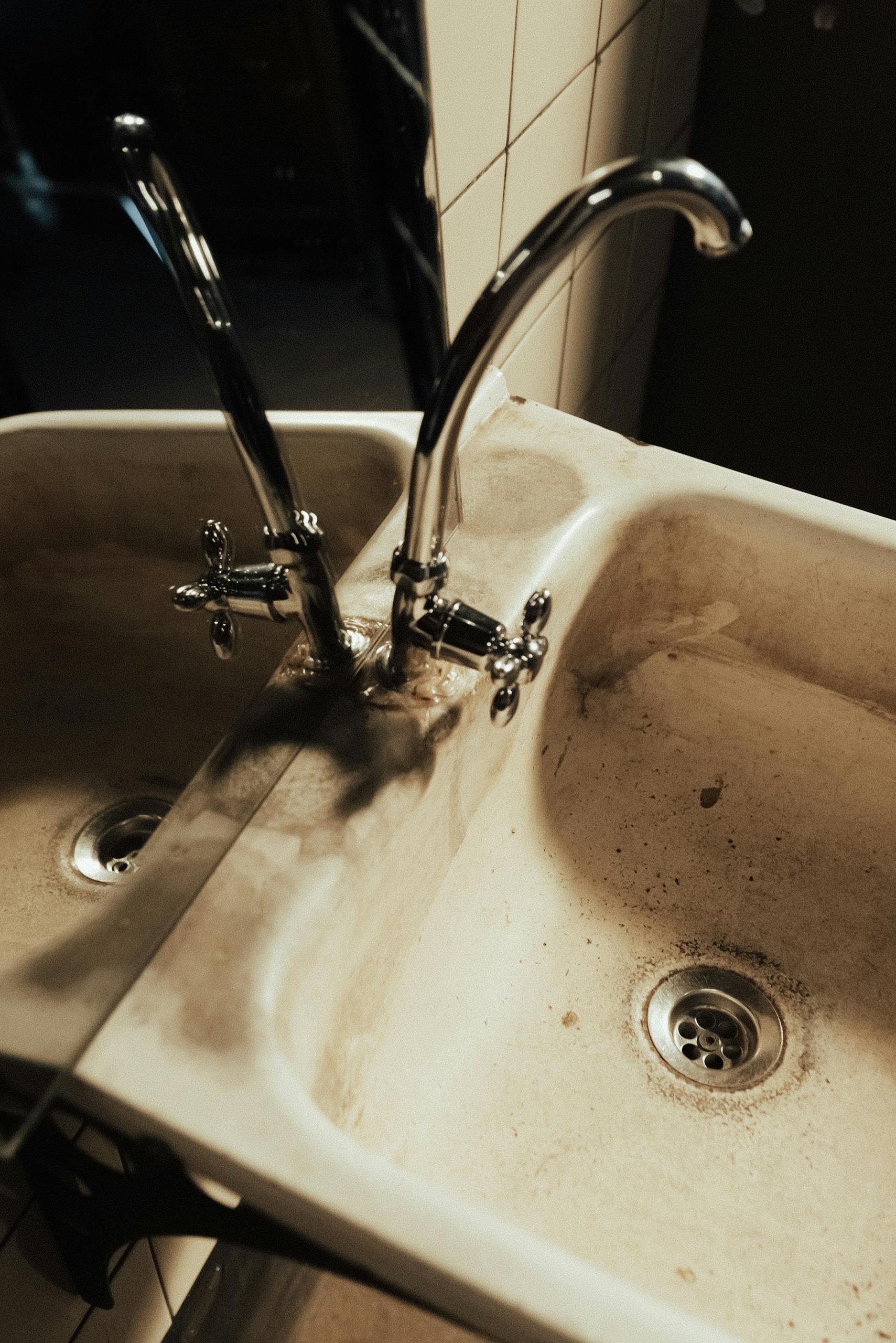 A neglected bathroom sink with visible dirt and a faucet reflection in the mirror.